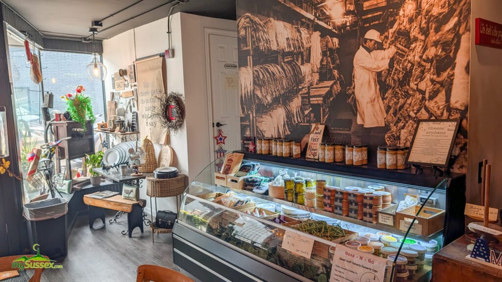 Cold case inside Curd & Cleaver in Newton, NJ, with cheeses and gourmet foods displayed beneath wallpaper showing a vintage butcher working with dry-aged beef.