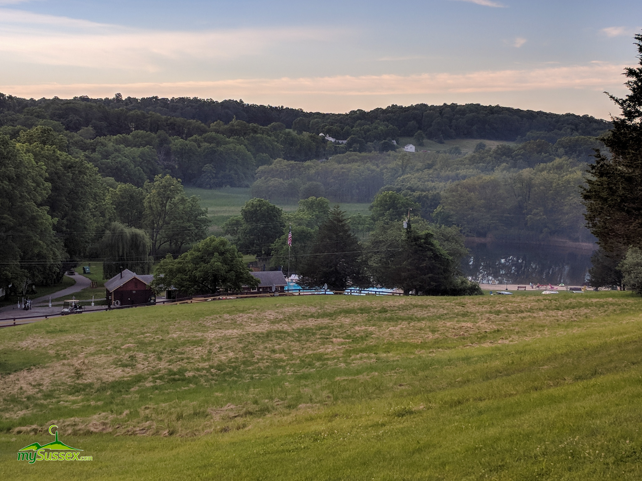 Scenic rolling landscape with green fields, forested hills, a river, and red barn buildings nestled below, typical of Sussex County's natural terrain